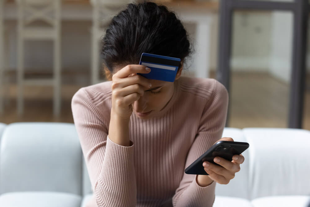 Upset latina female internet shopper sit on couch hold phone with opened web shop ebank page suffer of overspending money from card account. Frustrated young lady lost savings as scam operation result